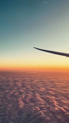 A vertical footage of the wing of a plane flying over the dense clouds at sunset, with a scenic dusk sky in the background