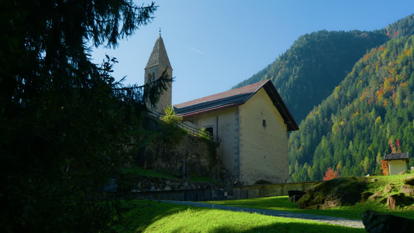 Wide shot of a small brick mountain church with bell tower and three crosses on a hill above a cemetery, next to a forest in nature.