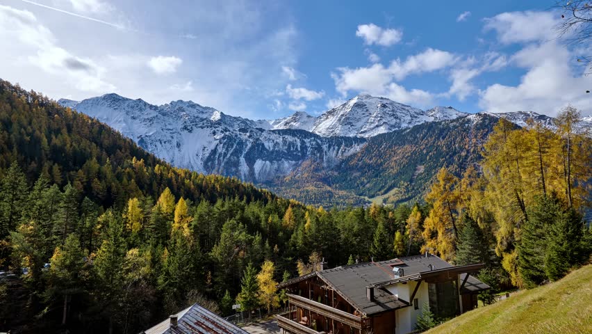 Autumn alpine valley with snowy mountains forest and chalet under partly cloudy blue sky timelapse