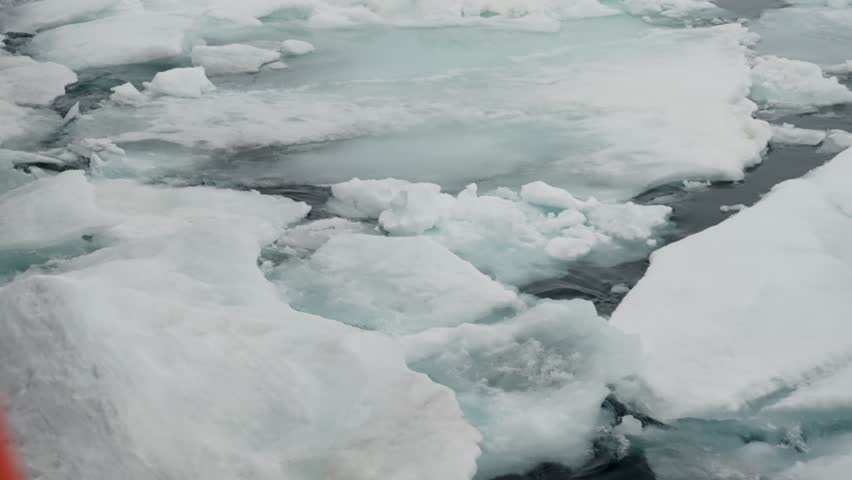 Drifting sea ice floes and fractured snow slabs on dark seawater, with turquoise meltwater pools and deep cracks, revealing layered textures across a cold polar seascape