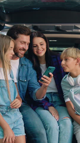 Cheerful lovely parents with cute teen kids which using mobile phone sitting in the car's trunk. Mother Father and daughter son surfing internet online on cellphone. Family road trip with trailer car