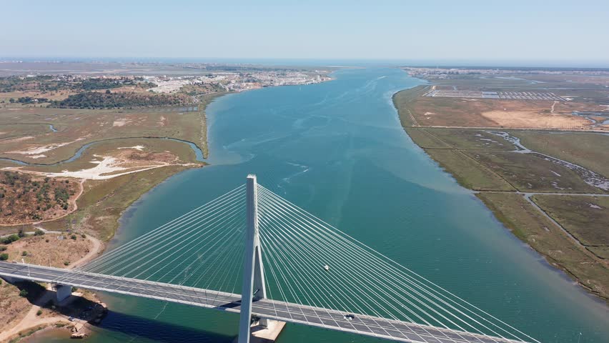 Ponte internacional Guadiana drone view of bridge and estuary, winding channel toward horizon, cable-stayed span in foreground and small boat leaving white wake, salt marshes and sandy