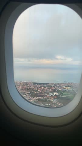 Aerial view of a coastal Porto, Portugal seen through an airplane window during soft sunrise light, revealing ocean, buildings, and winding waterways below.