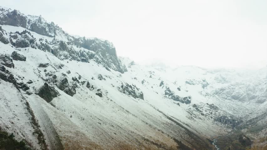 panoramic shot of the snow covered mountains in the North Caucasus region of Russia. The footage captures the rugged terrain, rocky slopes, and a hint of a stream running through the valley under a mo