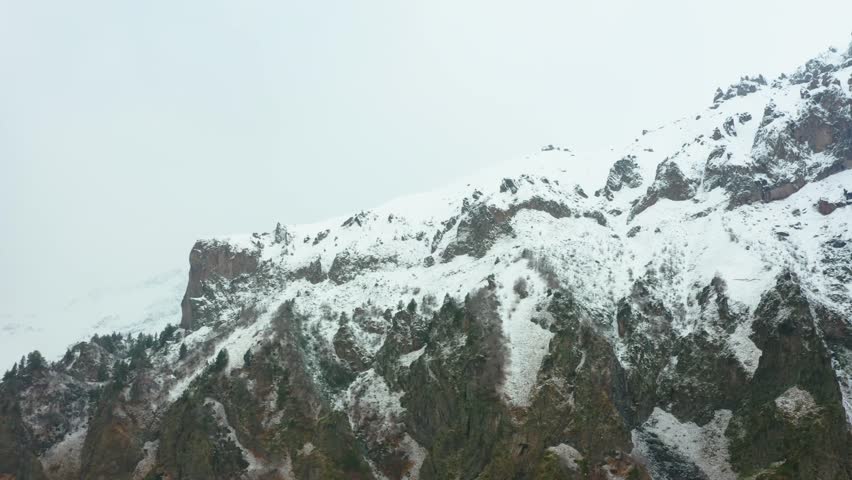 A scenic shot of the snow covered peaks and rocky terrain of the North Caucasus mountain range in Russia on an overcast day.