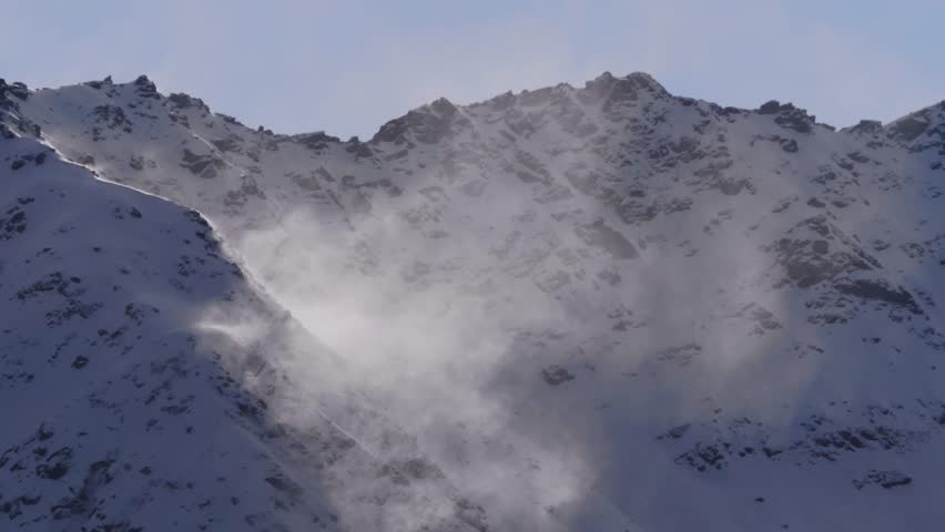A view of a snow covered mountain peak on a clear day, with gusts of wind blowing fresh powder across the rugged terrain. The bright sunlight illuminates the swirling snow, creating a dynamic and sere