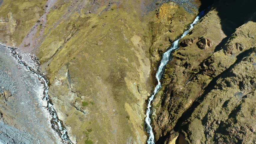 An aerial drone shot captures the stunning natural landscape of the North Caucasus mountains in Russia. A clear stream cascades down a rugged, grassy slope, highlighting the untouched beauty of the re