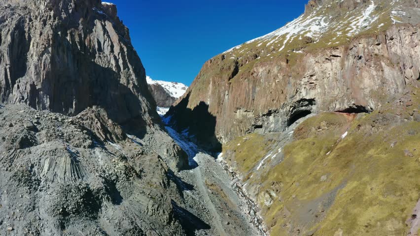 A aerial view of the North Caucasus mountain range in Russia, showcasing a rugged landscape with a prominent glacier flowing through a rocky valley.