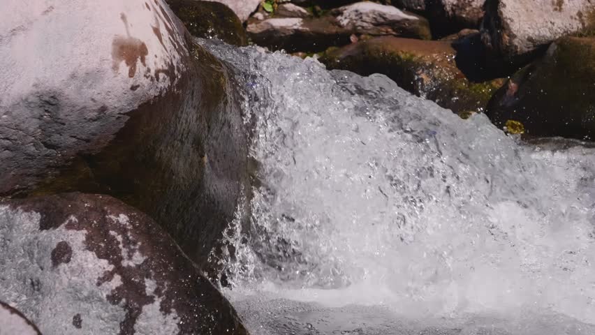 A close up shot of a small waterfall or cascade in a mountain stream. The clear, bubbling water rushes over smooth, wet rocks. The footage captures the movement of the water and the natural textures o