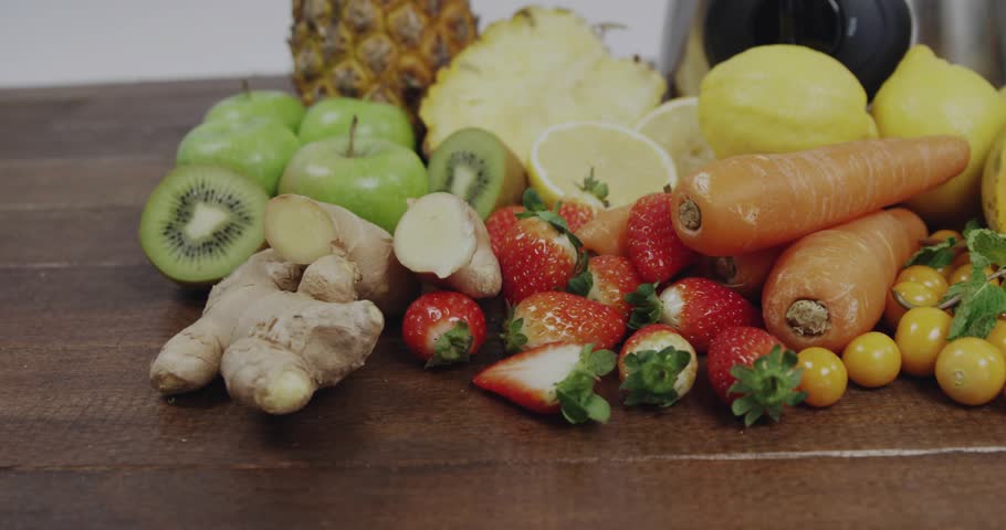 Camera pulling back revealing blender filling with sliced fruit for demo, binary overlay floating. Strawberries, pineapple, kiwi, papaya, carrots, ginger, countertop
