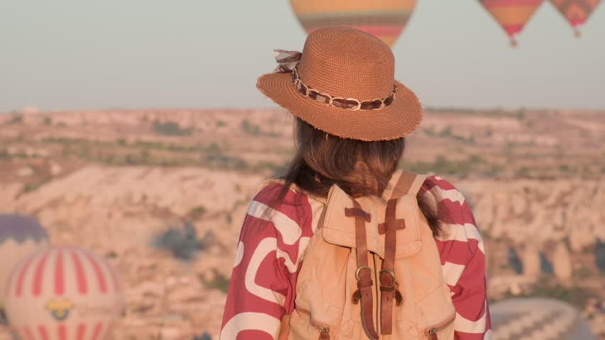 Inspired tourist woman in hat watching hot air balloons tour flying in sky in Cappadocia mountain valley at sunrise in morning