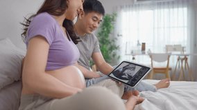 Asian young couple watching their baby's ultrasound on digital tablet. Attractive expectant husband and pregnant wife sitting on bed in bedroom, feels excited and enjoy look at image of unborn child. - Powered by Shutterstock - Get 15% off with code: PIKWIZARD15