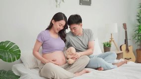 Asian expectant couple sharing loving moment, ready to become parents. Attractive pregnant wife and husband spend time together, placing teddy bear on belly while sitting on cozy bed in bedroom. - Powered by Shutterstock - Get 15% off with code: PIKWIZARD15