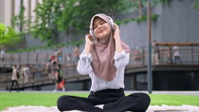 Asian Muslim woman enjoy music with headphones while sitting in the city. Attractive young girl wearing hijab feel happy and relax, having picnic and listen to songs outdoors in modern urban setting. - Powered by Shutterstock - Get 15% off with code: PIKWIZARD15