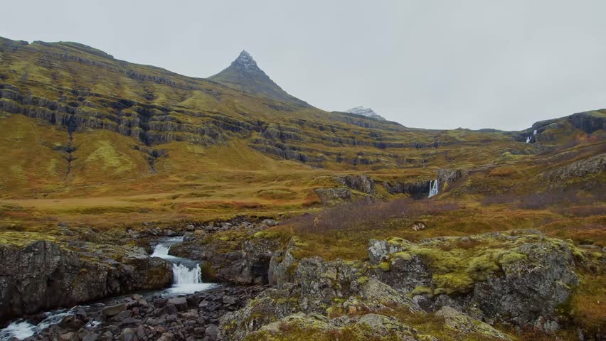 A stunning wide shot of a rugged Icelandic landscape. A small waterfall cascades over moss-covered rocks and into a stream, with a majestic, pointed mountain peak visible in the distance under an over
