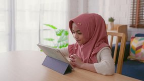 Asian Muslim girl using a digital tablet in cozy living room at home. Adorable young child feel happy and relax, studying at the table, attending virtual classes online and watching cartoons in house. - Powered by Shutterstock - Get 15% off with code: PIKWIZARD15