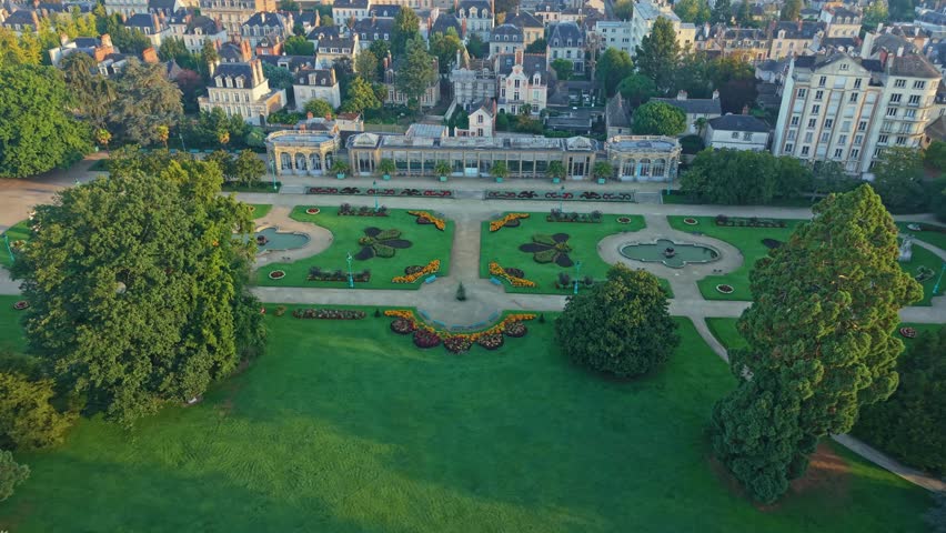 Drone shot descending toward Parc du Thabor in Rennes, revealing French-style gardens, paths, fountains, surrounding buildings and warm sunrise light.