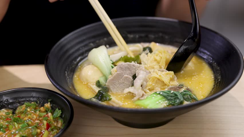 Hand uses chopsticks and spoon to eat spicy noodle soup with vegetables and chicken slices