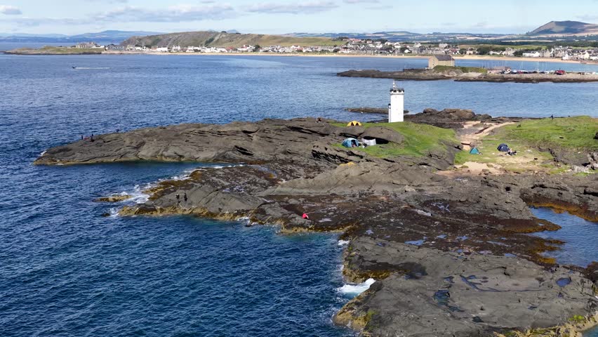 Drone pans over rocky shoreline, lighthouse, tents, and seaside village under bright daylight in Scotland