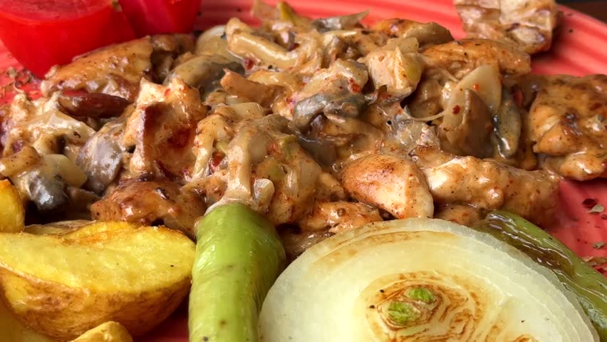 A vibrant plate featuring chicken and mushroom stroganoff served with golden potato wedges, a thick onion slice, a green chili, and a fresh tomato wedge.