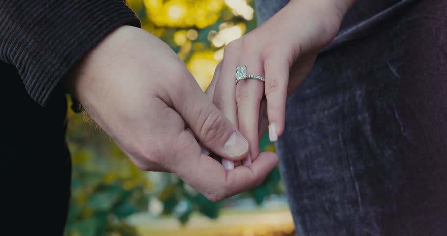 Close-up couple holding hands with ring, large diamond ring visible on woman's finger, man gently clasping hand, soft golden hour
