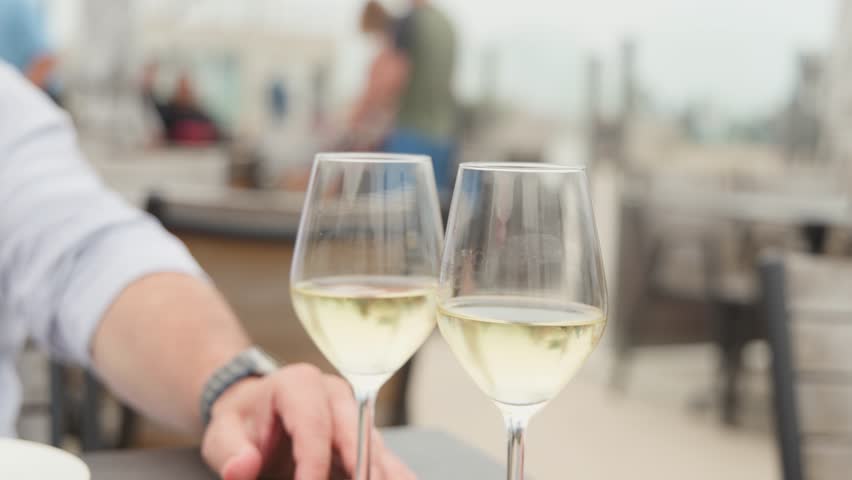 Person clinks white wine glasses outdoors at coastal cafÃ©, soft daylight, shallow depth of field