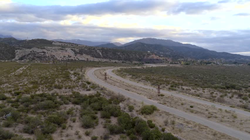 A winding paved road cuts through a rugged valley, mountain ridges in the distance, under a cloud-filled sky near Estación 2.