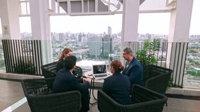 Two business professionals shake hands outdoors on a rooftop, while colleagues sit nearby with a laptop. The group engages in a collaborative meeting with a city skyline in the background. 4k video - Powered by Shutterstock - Get 15% off with code: PIKWIZARD15