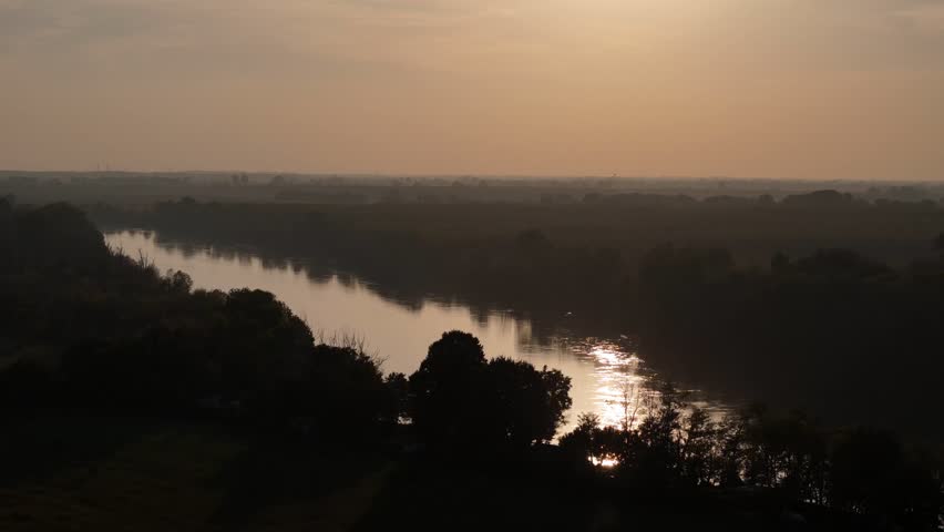 Wide shot captures Po River and wetlands of Isola Serafini with golden haze at sunset near Olza, Monticelli d’Ongina, Piacenza, Emilia-Romagna, Italy with, calm waters, woodland silhouettes.