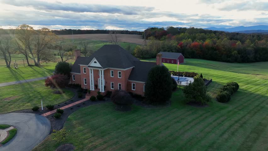 Aerial orbit shot of luxurious countryside estate with large mansion, detached garage, driveway, manicured lawn and backyard pool. Surrounded by colored forest trees in autumn. Virginia, USA.