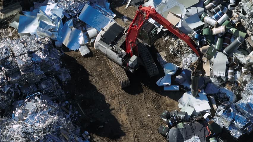 Working Excavator on scrapyard in industrial area of town. Aerial top down flyover. Sunny day. steel and aluminum on junkyard.