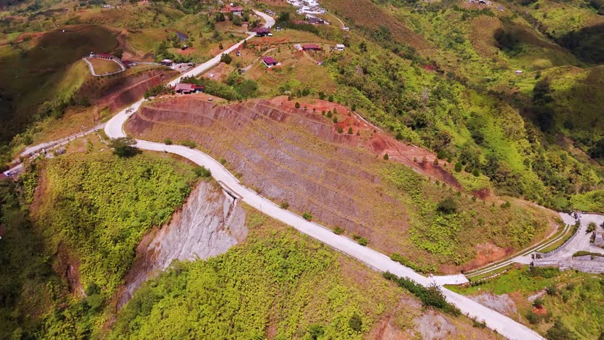 Aerial 4K footage showing a winding mountain road, hillside homes, terraced slopes, and a dramatic cliff drop surrounded by lush green highland terrain.