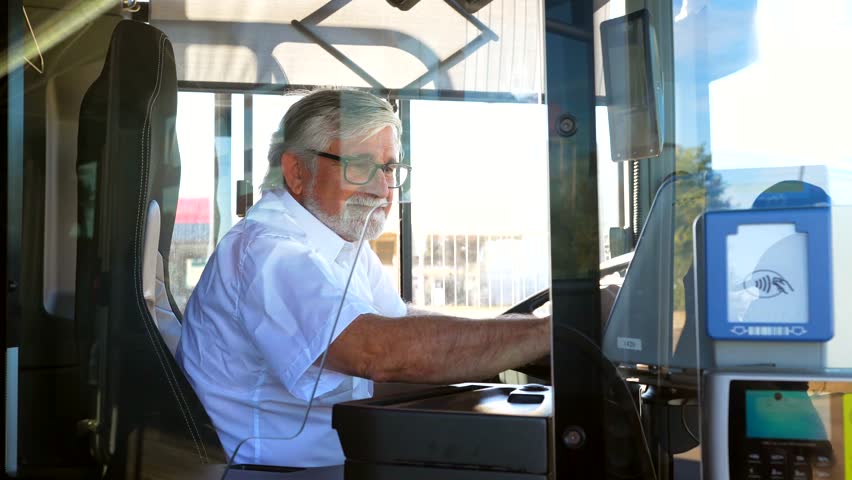 Experienced senior male bus driver with a beard and glasses maneuvering the steering wheel while driving, glancing back at the camera and smiling, illustrating a public transport career
