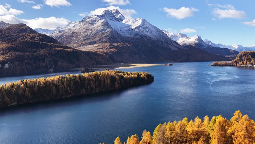 Snow-capped alpine mountains with golden larch forest and pristine blue lake waters.