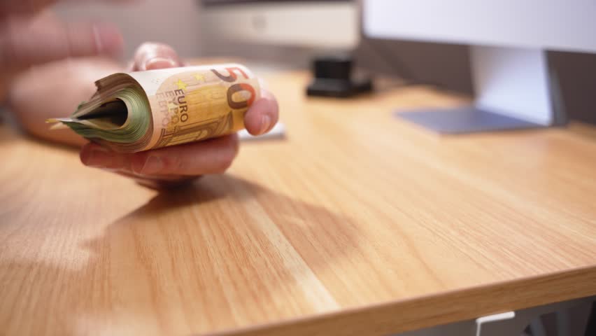 Hand presents a rolled bundle of euro banknotes over a wooden desk. The clip visualises cash payout, savings roll or secret stash of money.