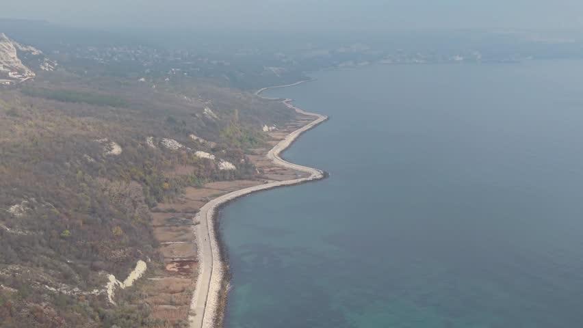 Aerial drone video showing winding coastal road beside calm sea, capturing natural shoreline landscape, rugged terrain, and peaceful seaside scenery from above.