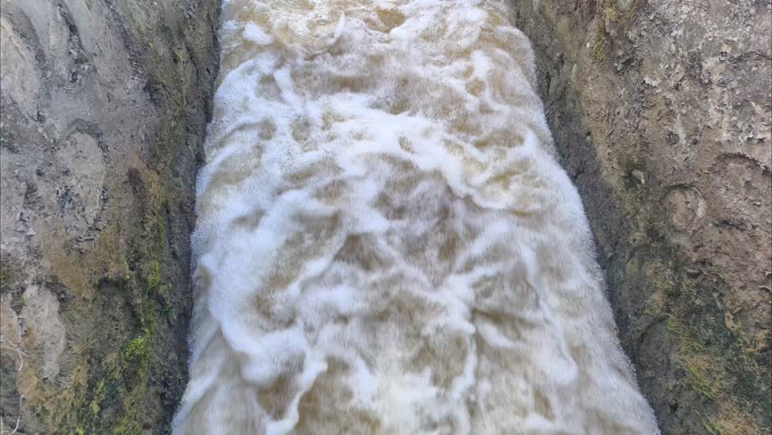 A photo of water flowing rapidly through narrow man-made channels, such as drainage or irrigation systems. The water appears foamy and murky, flowing rapidly, possibly containing mud or sediment. 