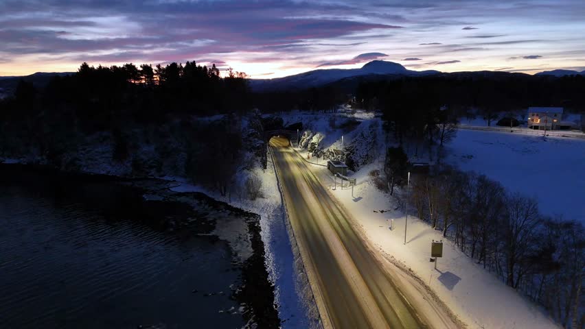 A drone footage of cars driving on the snow-covered Riksvei 70 highway, between sea and line of trees at dawn in Kristiansund, Norway