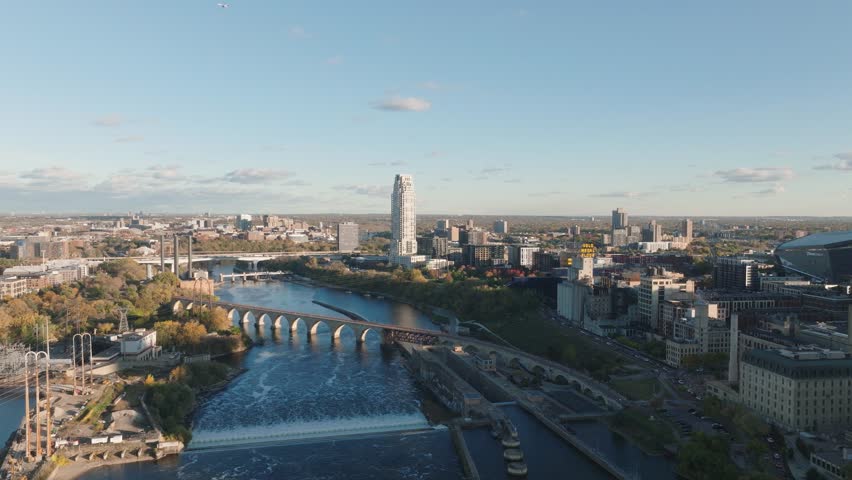 Aerial descent over Mississippi River, Minneapolis. Features Stone Arch Bridge, St. Anthony Falls Dam, and Downtown Skyline with the Eleven Tower and Gold Medal Flour sign at sunset golden hour.