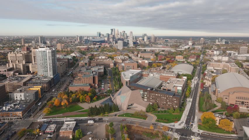 Wide aerial drone shot over the U of M East Bank campus during fall. Features the McNamara Center, Williams Arena, and the Minneapolis skyline background.