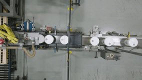 Top-down aerial shot of a yellow robotic arm performing palletizing by stacking white pails or buckets onto wooden pallets on an automated conveyor belt in a manufacturing factory. - Powered by Shutterstock - Get 15% off with code: PIKWIZARD15