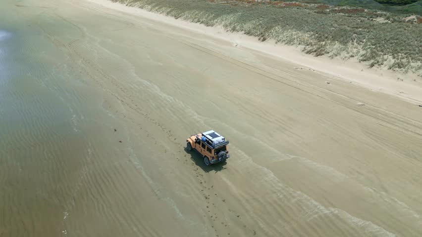 A Drone footage of a 4WD adventure car driving on a sandy beach along blue calm sea water