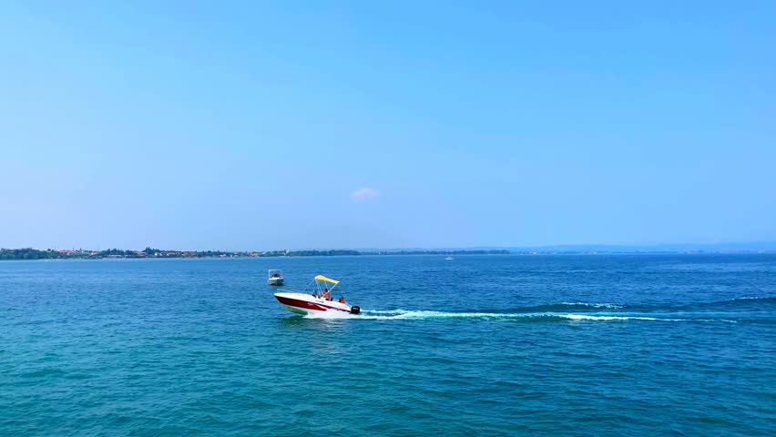 Hectic scene from Lago di Garda with different motorboats, stationary and cruising the lake, leaving wakes on the wavy surface of the waters, with a vision of distant thickets, buildings and mountains