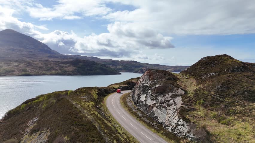 A drone footage of NC500 road through west coast of Scotland, surrounded by water and rugged terrain under cloudy sky
