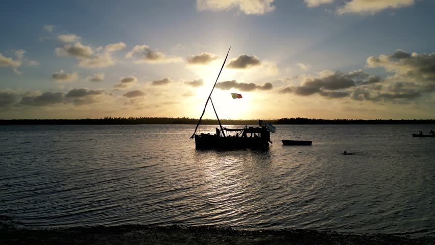 A drone flying above silhouetted traditional boat with flags, revealing the expanse of the calm sea and silhouetted shore in the background at sunset