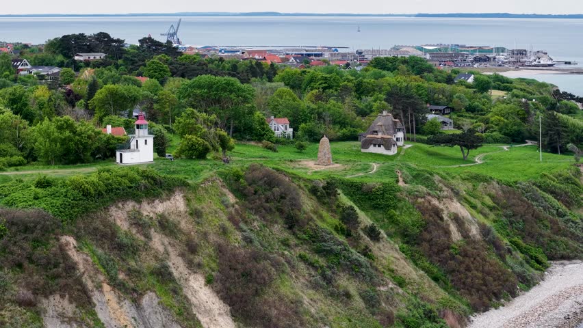 Drone footage of lush green hills and steep coastal cliffs beside the sea, featuring small houses and a lighthouse near Ega and Mols Bjerge in Denmark.