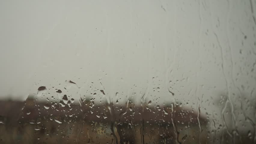 Raindrops cascade down the window, blurring the view of the peaceful landscape outside. This early evening rain brings a sense of tranquility to the surroundings. Dramatic sky.