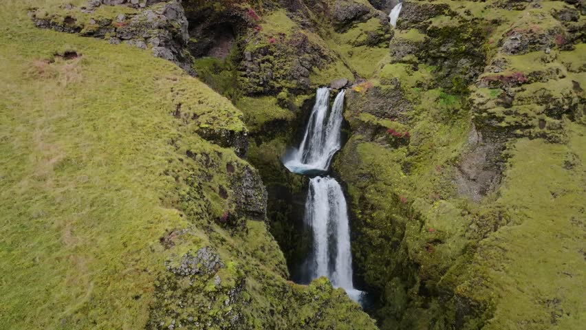 Aerial view of a striking two-tiered waterfall cascading down a steep, moss-covered canyon, creating a mesmerizing display of nature's power, Iceland.