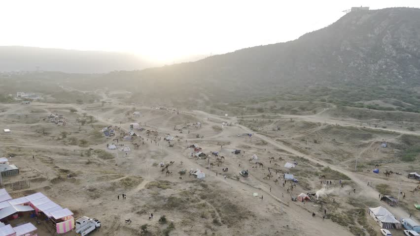 Aerial shot of Pushkar cattle fair ground during sunrise, showing camels and tents with the Aravalli mountains in the background under warm morning light.