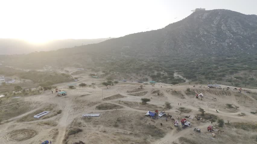 Aerial shot of Pushkar cattle fair ground during sunrise, showing camels and tents with the Aravalli mountains in the background under warm morning light.
